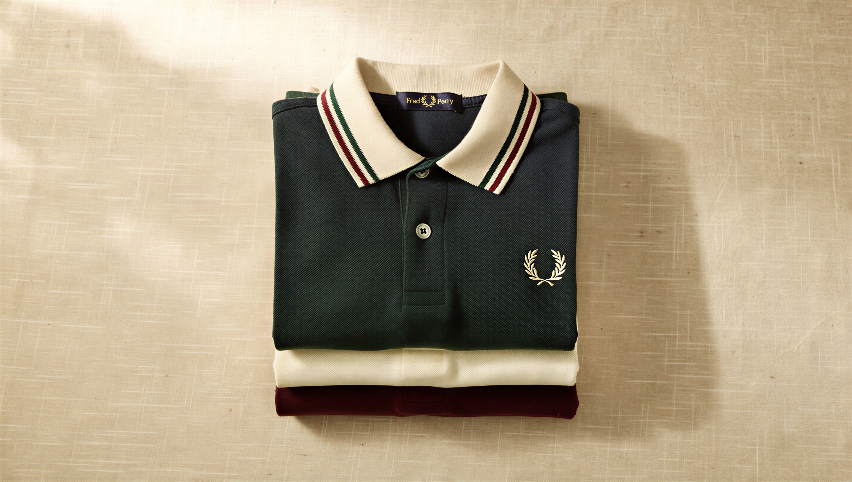 Ein Stapel gefalteter Fred-Perry-Polos in verschiedenen Farben auf cremefarbenem Leinen — ein Reseller-Dauerbrenner, der sich über das ganze Jahr verlässlich verkauft.